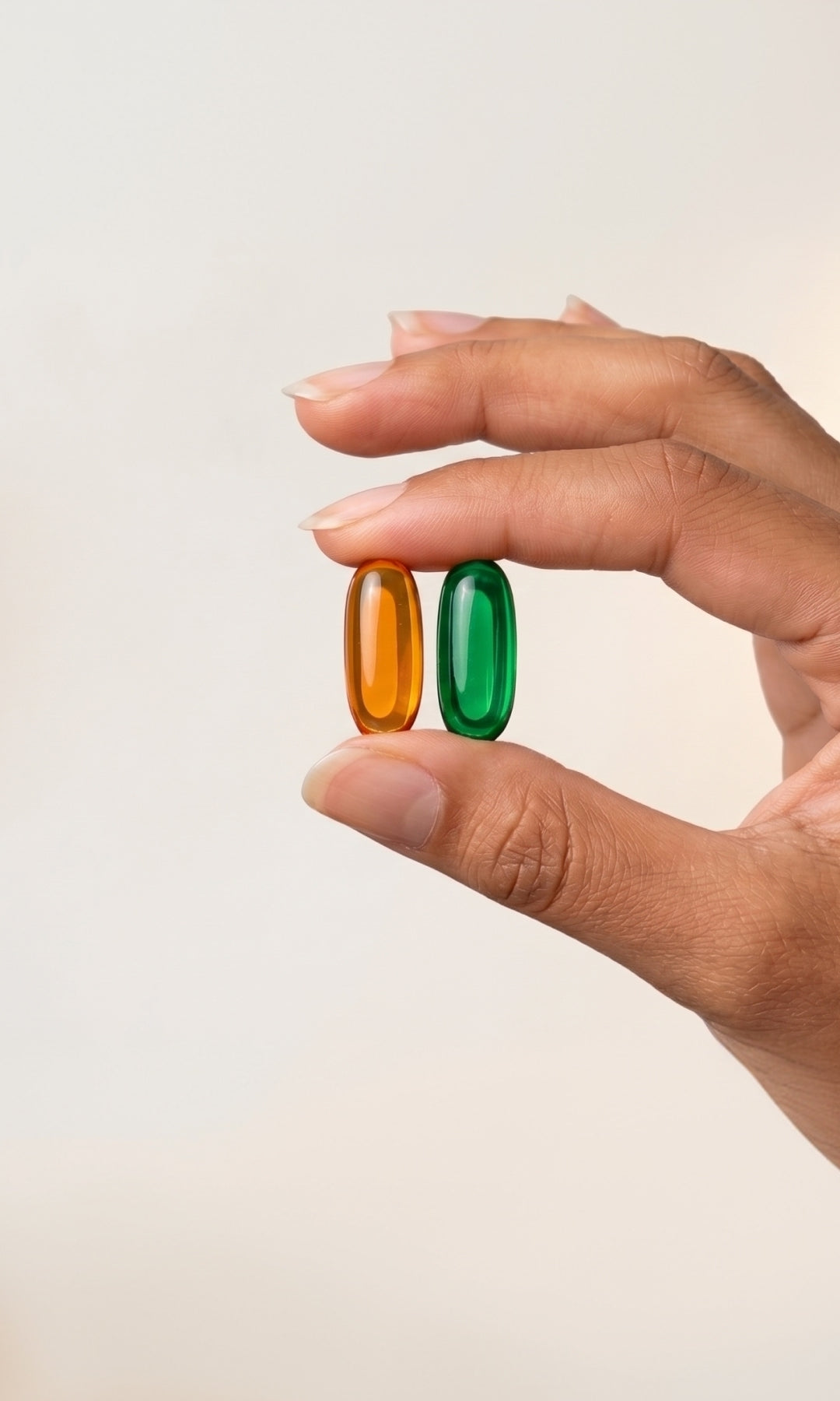 A close-up of a hand holding two translucent capsules, one amber-orange and one green, against a plain light beige background.