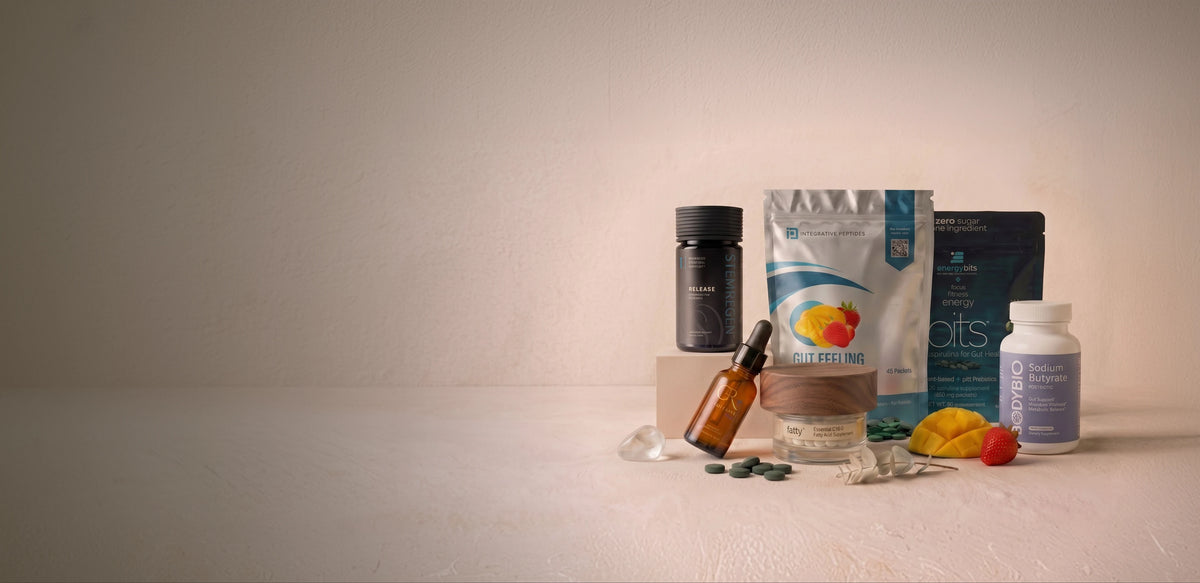 A group of dietary supplement products arranged on a light beige surface against a matching neutral background. The products include a silver and white pouch with a yellow graphic, a dark blue bag, a white bottle, a black bottle, a brown glass dropper bottle, loose green capsules, and small yellow and red fruit pieces.